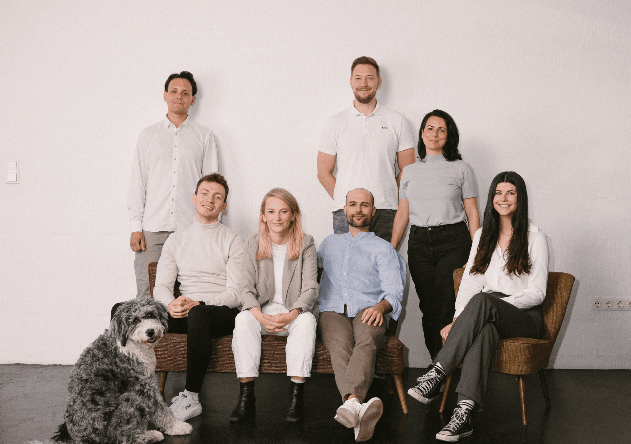 A group of four casually dressed people posing in a bright, modern room with large windows and bookshelves. They are looking confidently at the camera, with natural light filling the space.